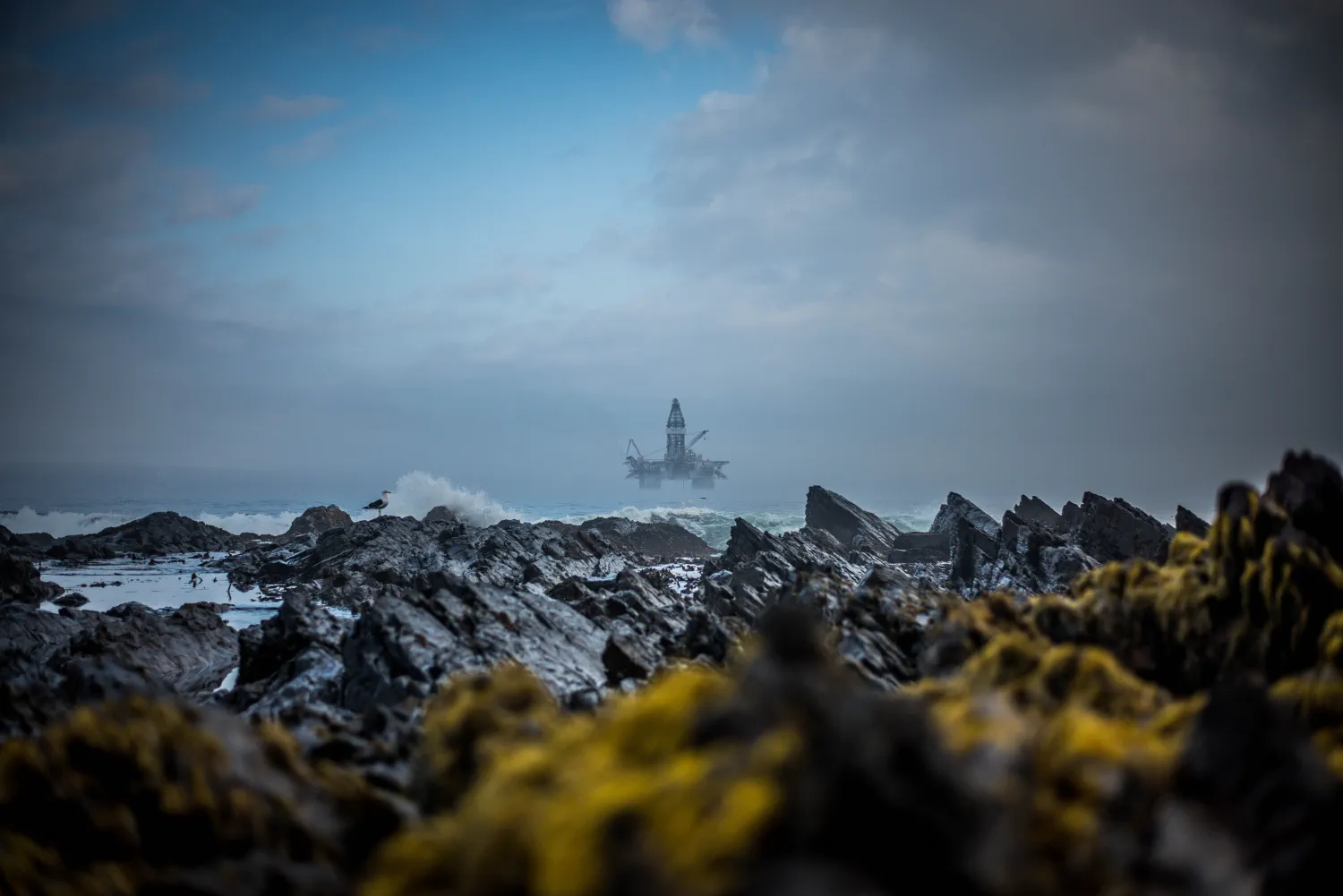 Waves breaking in harsh terrain, with offshore oil rig visible in a distance