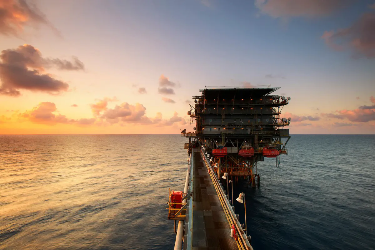 Offshore oil platform at sunset, connected by a metal walkway extending over calm ocean waters.